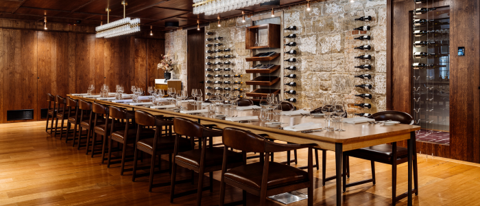 Tasman (Eat & Drink) 2 An elegant dining room featuring a long wooden table set for a meal with plates, glasses, and cutlery. Wooden chairs surround the table, and the room has wooden floors and walls. One wall is lined with wine racks filled with bottles, and modern light fixtures hang from the ceiling.