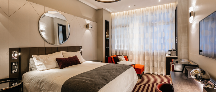 Tasman (Stay) 2 A stylish hotel room with a large bed dressed in white linens and a dark gray throw. A round mirror hangs above the bed, flanked by two bedside tables with lamps. The room includes a window with sheer curtains, two orange chairs, and a small round table. A flat-screen TV is mounted on the wall above a desk with an upholstered chair. The floor features a striped carpet in brown and beige tones.