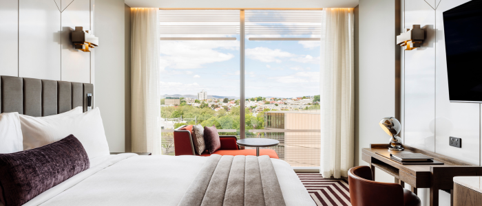 Tasman (Stay) 3 A modern hotel room with a large bed dressed in white linens, a dark gray blanket, and a dark red bolster pillow. A round mirror hangs above the bed, with wall-mounted reading lights and a bedside table holding an alarm clock and phone. An orange armchair with an ottoman sits by a window with sheer curtains. The room features striped brown and beige carpeting, and a flat-screen TV mounted above a desk area.