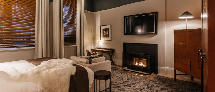 Tasman (Stay) 4 A cozy, modern bedroom featuring a bed with white and brown bedding, a small side table, and an armchair. A lit fireplace with a flat-screen TV mounted above it adds warmth to the room. To the right of the fireplace is a wooden cabinet with a lamp. A desk and chair sit in the corner beneath a piece of wall art. Large windows with wooden blinds and white curtains let in natural light.
