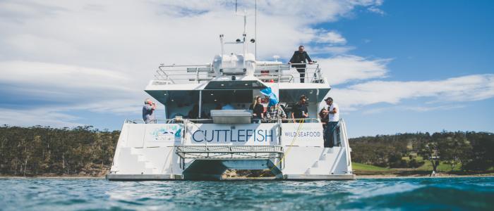 A large white boat named "Cuttlefish Wild Seafood" is anchored on a body of water. Several people are standing on the deck, some looking out at the water and others engaged in various activities. The boat has multiple levels and equipment visible on top. In the background, there are trees and a grassy shoreline under a partly cloudy sky