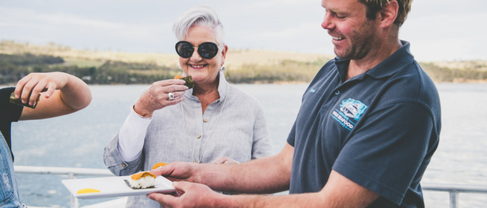 Three people are on a boat or near water. One person is holding a plate with freshly caught seafood and offering it to another person who is about to eat it. The background includes water and a distant shoreline.