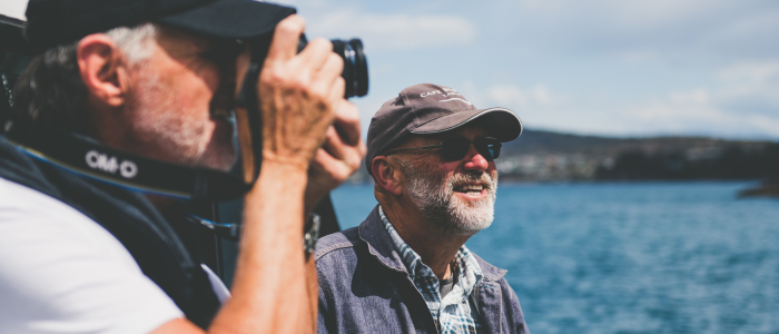 Two individuals are outdoors near a body of water. One person is holding a camera up to their face, appearing to take a photo. The other, wears a hat and casual clothing. The background features water and distant hills under a partly cloudy sky