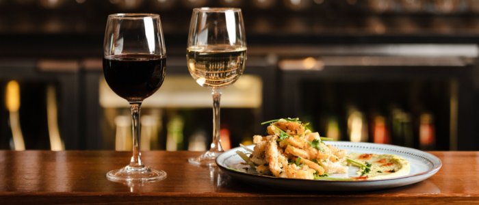 Two glasses of wine—one red and one white—placed on a wooden surface, with a plate of what looks like seafood and garnishes in front. The background shows a bar or restaurant setting with shelves and bottles visible.