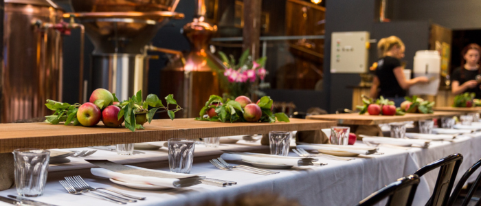 Long dining table set with white tablecloths, glassware, and neatly arranged cutlery, featuring wooden centerpieces decorated with fresh apples and green foliage, in a room with large copper distilling equipment in the background.