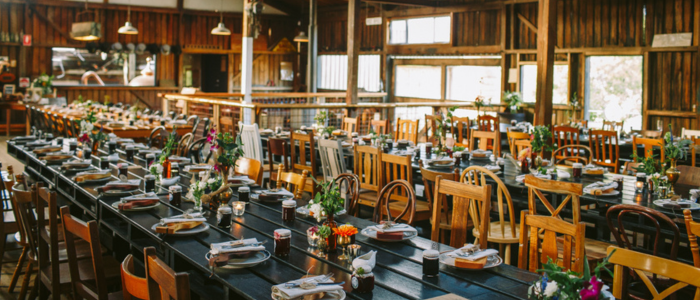 Rustic indoor dining space with long wooden tables set for a meal, featuring plates, cutlery, glassware, and small floral centerpieces, surrounded by a mix of wooden chairs in a warm, timber-paneled room with large windows.