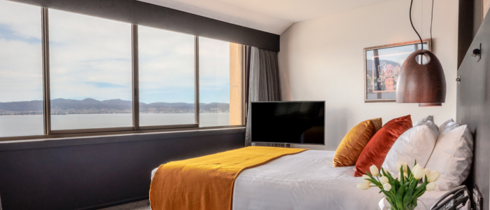 Wrest Point 4 Stay 2025 Stylish hotel bedroom with a large window overlooking water and hills. The room features a bed with white linens, orange accents, a wall-mounted TV, pendant light, bedside table with white tulips, and framed artwork.