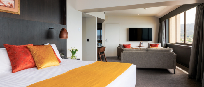 Wrest Point 5 Stay 2025 Modern hotel room with a large bed featuring white linens, an orange throw, and decorative pillows. The room includes a sitting area with a grey sofa, orange cushions, a round table with chairs, a wall-mounted TV, and large windows offering natural light and scenic views.