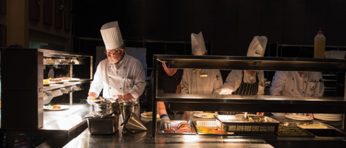 Wrest Point Eat&Drink 1 Group of chefs in white uniforms and hats working in a professional kitchen at Wrest Point Hotel & Conference Centre. They are preparing and plating food under bright lights on a stainless steel counter, with various kitchen utensils and equipment visible. The dark background contrasts with the well-lit workspace.