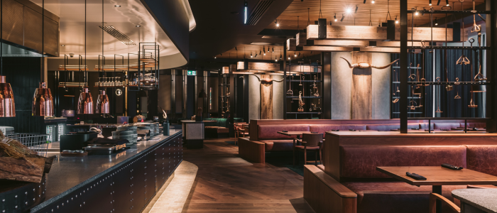 Wrest Point Eat&Drink 2 Interior of an upscale restaurant at Wrest Point Hotel & Conference Centre featuring brown leather booths, wooden tables, and a warm-toned design. The ceiling has wooden beams with hanging lights, and an open kitchen area with copper pots and pans is visible on the left. Decorative longhorn skulls adorn the walls, contributing to the sophisticated ambiance.