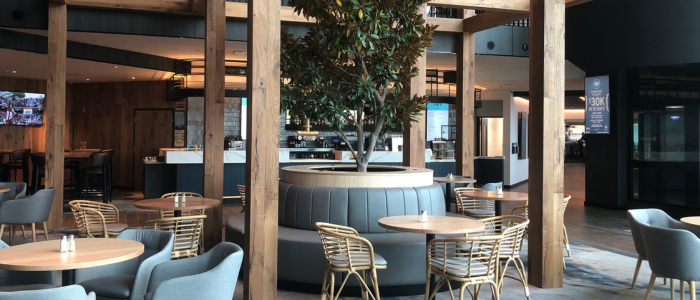 Wrest Point Eat&Drink 3 Interior of a modern café or restaurant at Wrest Point Hotel & Conference Centre featuring wooden beams, a central potted tree with cushioned seating, round tables with wicker and cushioned chairs, and a bar area with stools.