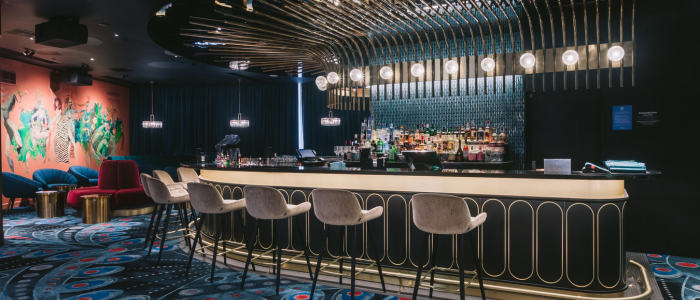Wrest Point Eat&Drink 4 Stylish bar at Wrest Point Hotel & Conference Centre featuring a curved counter with gold accents, high chairs, and a display of bottles behind the bar. The ceiling has an artistic light arrangement, the floor is covered with patterned carpet, and colorful abstract wall art is visible on the left.