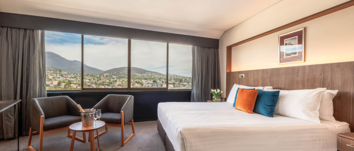 Wrest Point Stay 1 2025 Bright hotel room with a large window overlooking a mountain and town view. The room includes a neatly made bed with white linens and colorful pillows, two armchairs, a round table with an ice bucket and glasses, and a framed picture above the bed.