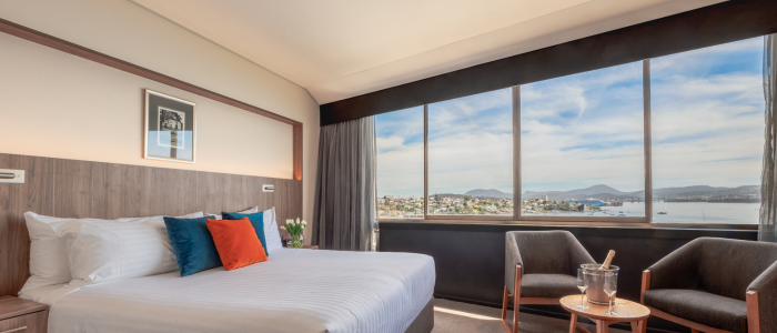 Wrest Point Stay 2 2025 Bright hotel room with a large window overlooking a mountain and town view. The room includes a neatly made bed with white linens and colorful pillows, two armchairs, a round table with an ice bucket and glasses, and a framed picture above the bed.