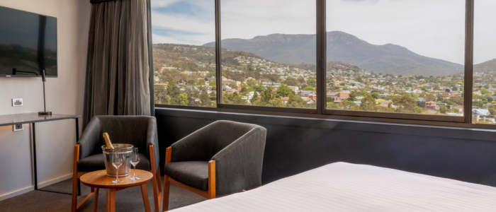 Wrest Point Stay 3 2025 Hotel room with a large window showcasing a mountain and residential view. The room includes a bed with white linens, two gray armchairs around a small table with an ice bucket and wine glasses, and a wall-mounted flat-screen TV.